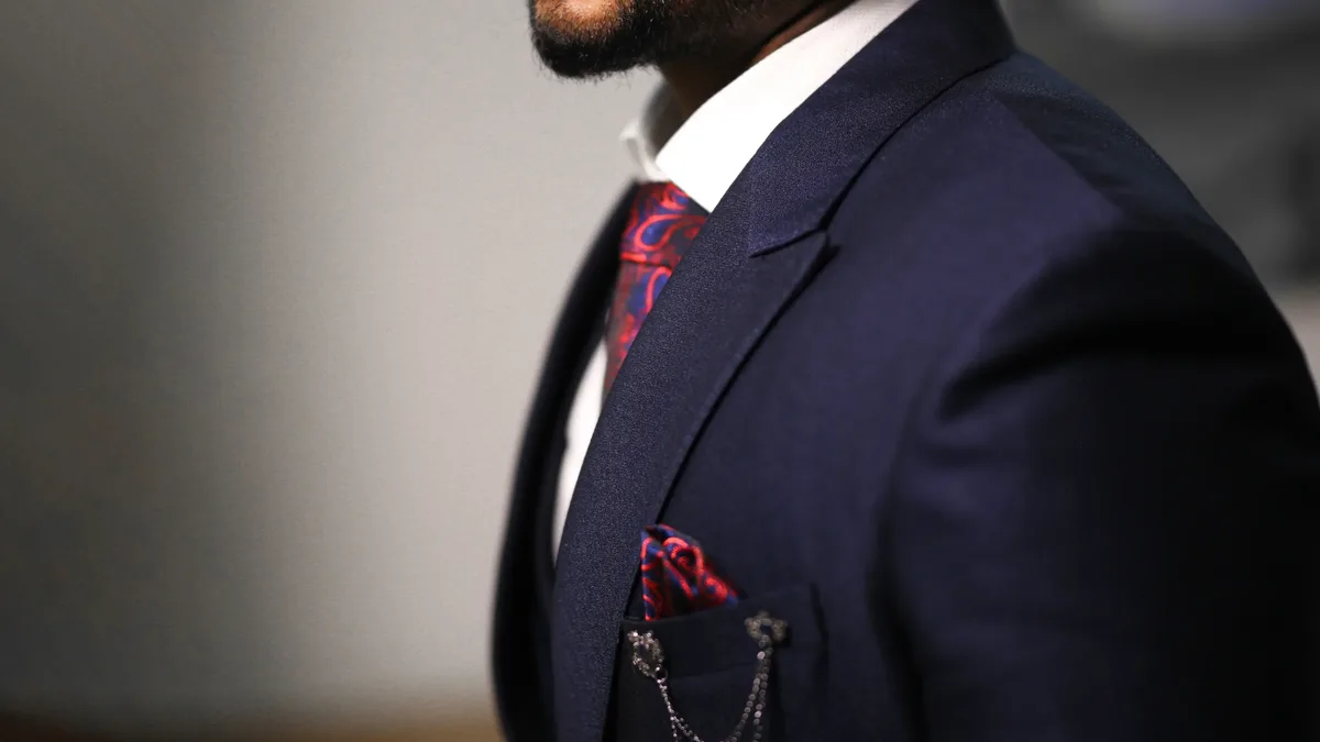 A man's torso, wearing a dark blue suit jacket, white shirt, red tie, and red pocket square with a silver chain attached to it.